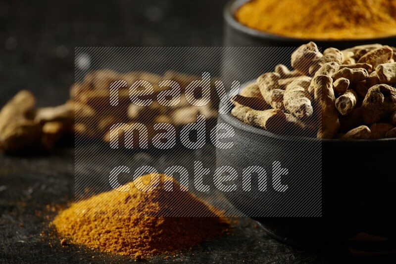 2 pottery black bowls, one full of turmeric powder and the other full of dried turmeric whole fingers on a textured black flooring