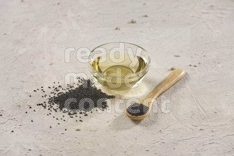 A glass bowl full of black seeds oil and wooden spoon full of black seeds with seeds spread on a textured white flooring