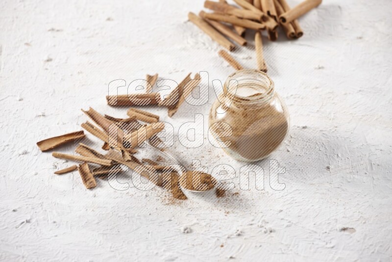 Herbal glass jar full cinnamon powder and a metal spoon surrounded by cinnamon sticks on a white background