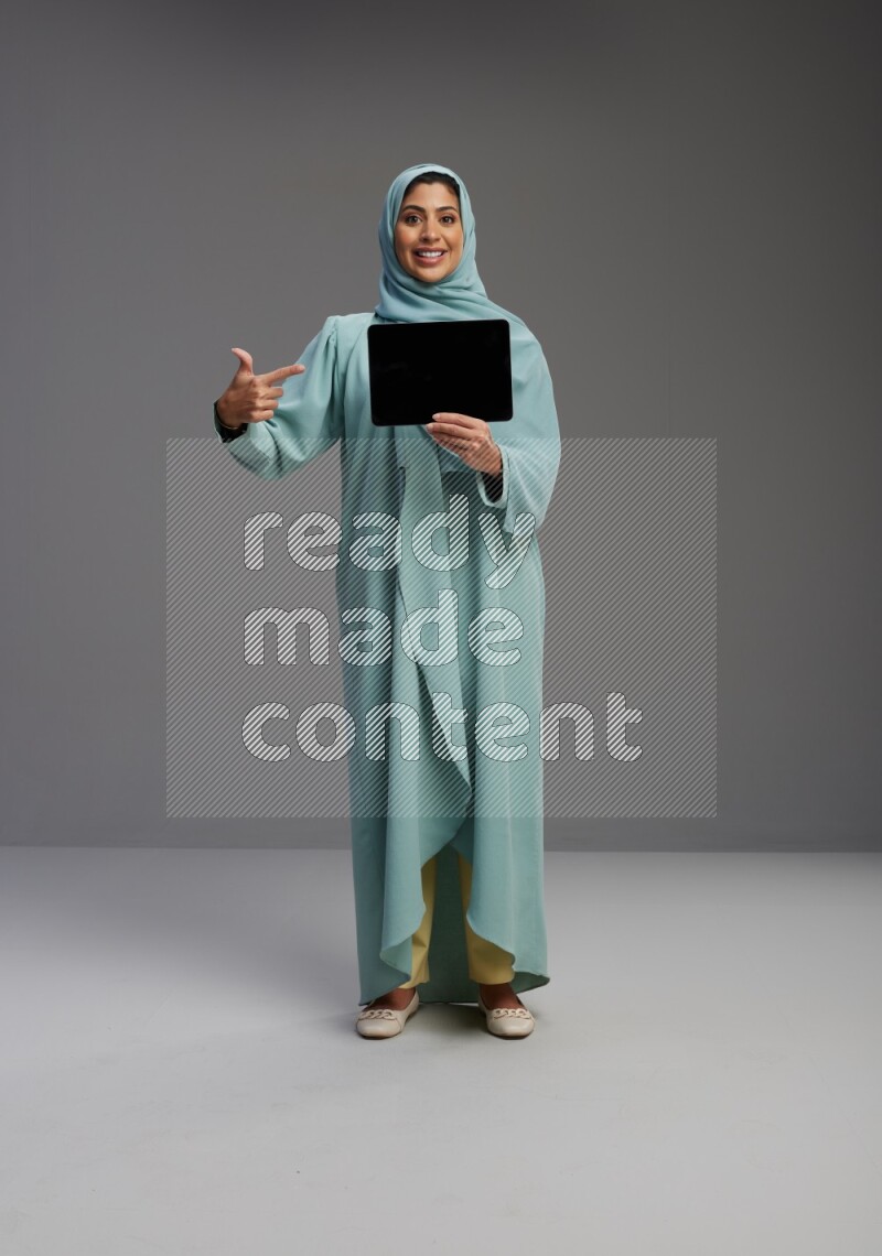 Saudi Woman wearing Abaya standing showing tablet to camera on Gray background