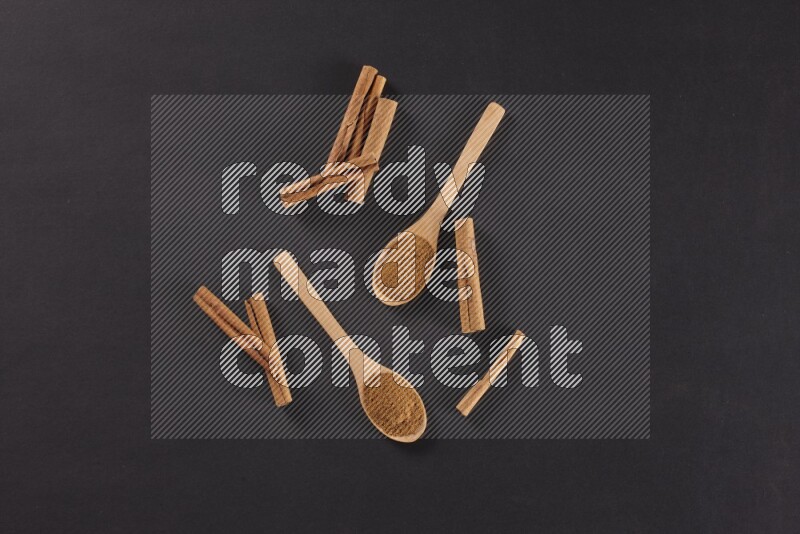 Two wooden spoons full of cinnamon powder with cinnamon sticks on black background
