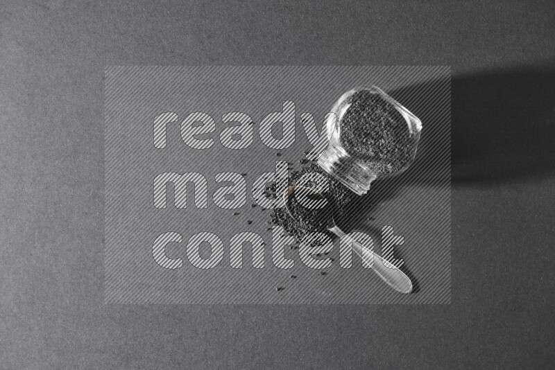 A flipped glass spice jar full of black seeds and the seeds spread out with a metal spoon full it on a black flooring