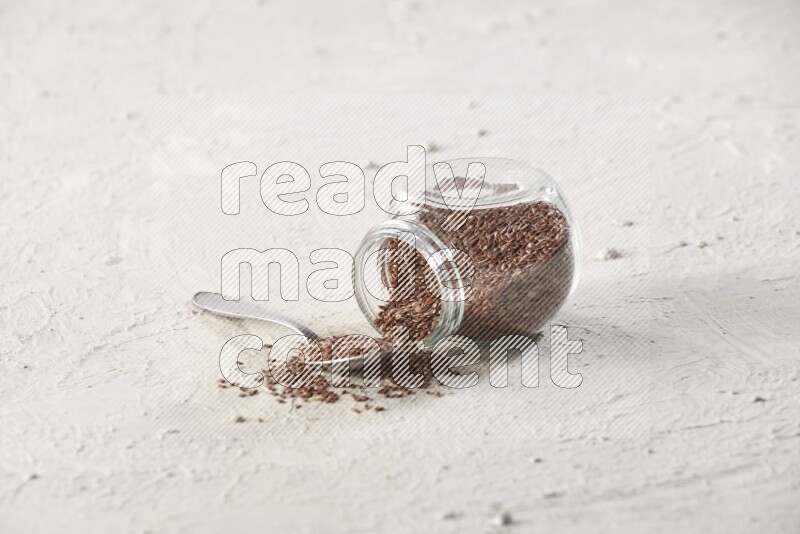 A glass spice jar full of flax seeds flipped with a metal spoon full of the seeds on a textured white flooring