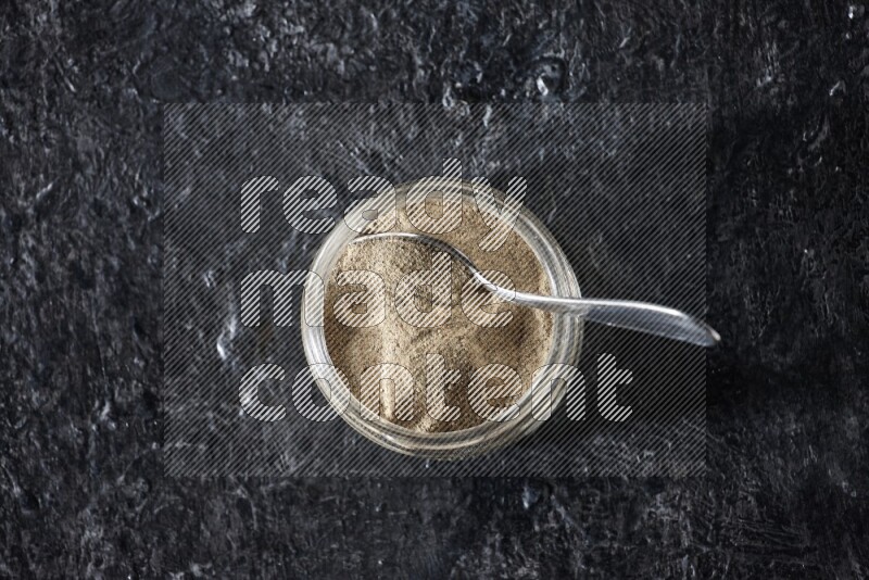 A glass jar and a metal spoon full of white pepper powder on textured black flooring