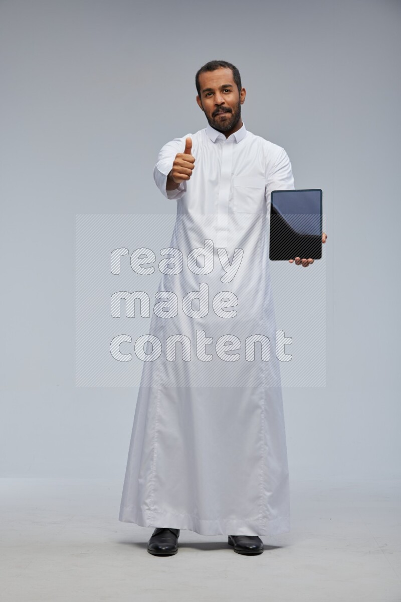 Saudi man wearing Thob standing showing tablet to camera on Gray background