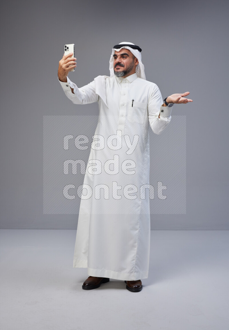 Saudi man Wearing Thob and white Shomag standing taking selfie on Gray background