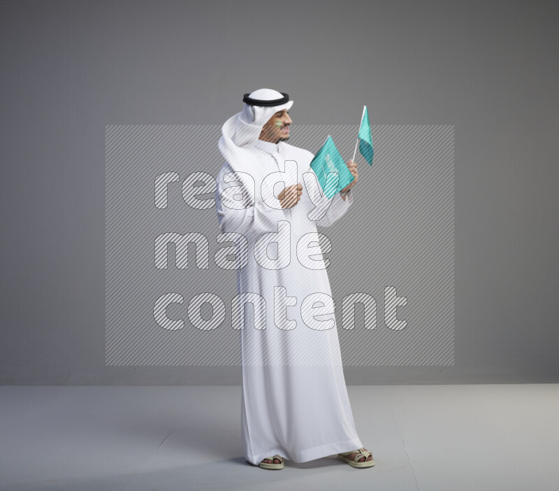 A Saudi man standing wearing thob and white shomag with face painting raising small saudi flag on gray background