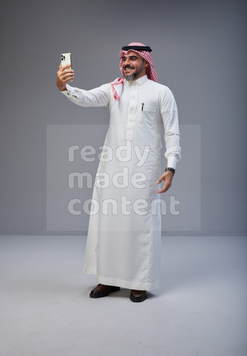 Saudi man Wearing Thob and red Shomag standing taking selfie on Gray background