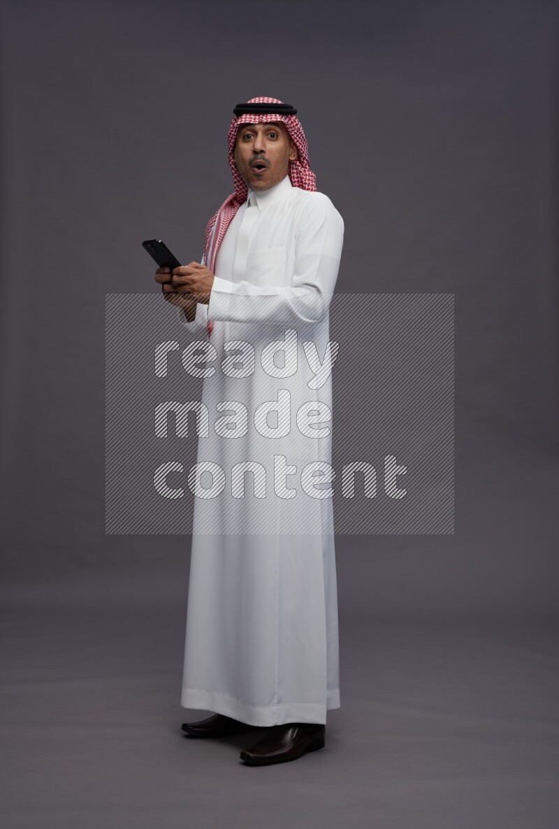 Saudi man wearing thob and shomag standing texting on phone on gray background