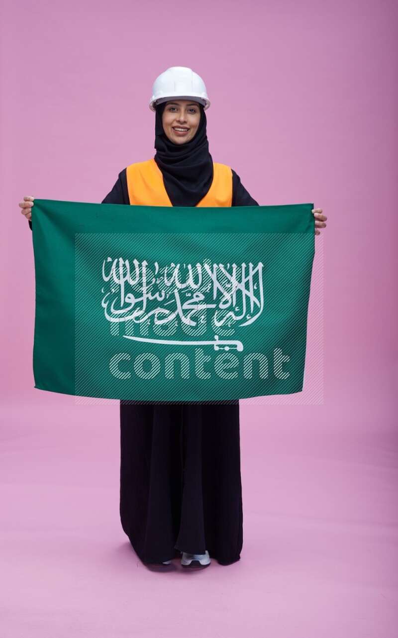 Saudi woman wearing Abaya with engineer vest and helmet standing holding Saudi flag on pink background