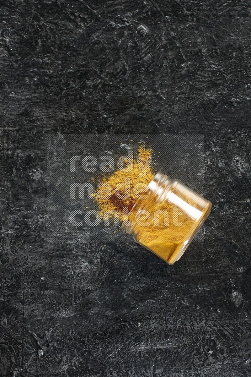 A flipped glass jar full of turmeric powder and powder spilled out of it on a textured black flooring
