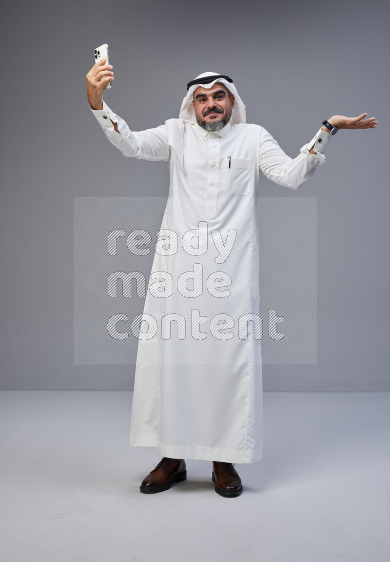 Saudi man Wearing Thob and white Shomag standing taking selfie on Gray background