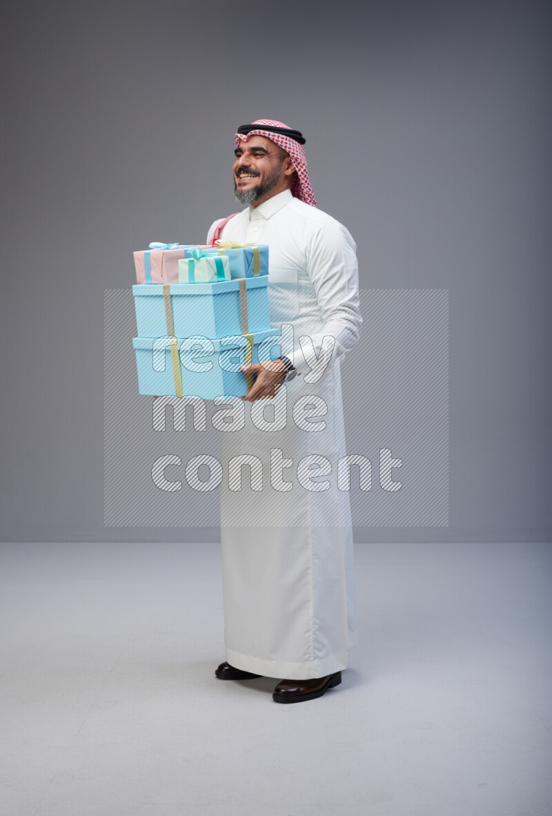 Saudi man Wearing Thob and red Shomag standing holding gift box on Gray background