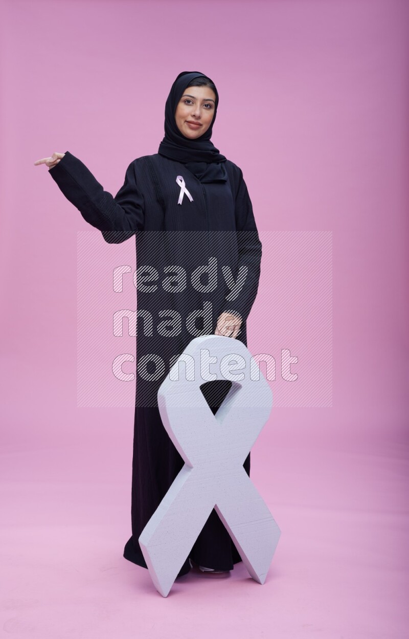 Saudi woman wearing pink ribbon on abaya standing holding awareness ribbon on plain pink background