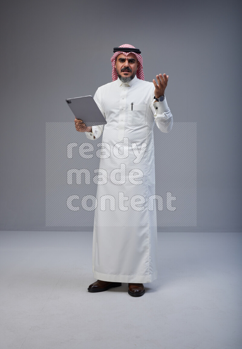 Saudi man Wearing Thob and red Shomag standing working on tablet on Gray background