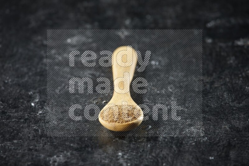 A wooden spoons full of cumin powder on textured black flooring
