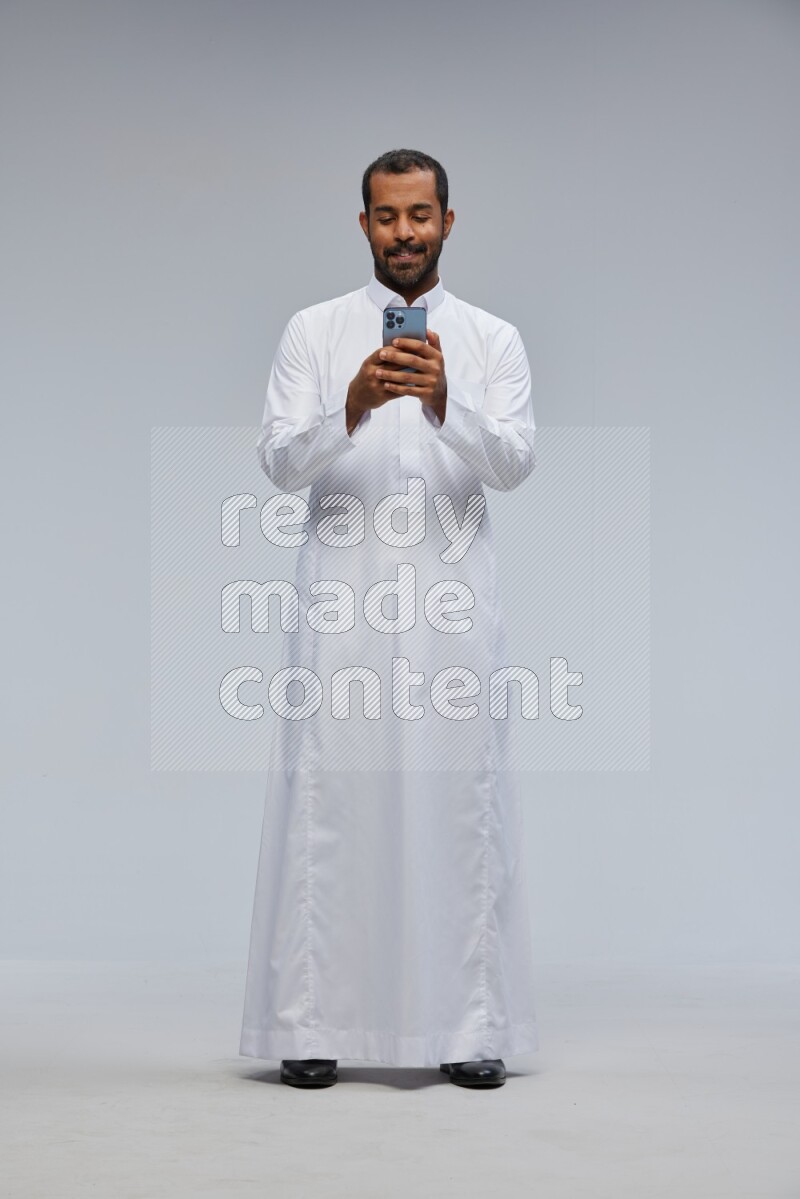Saudi man wearing Thob standing texting on phone on Gray background
