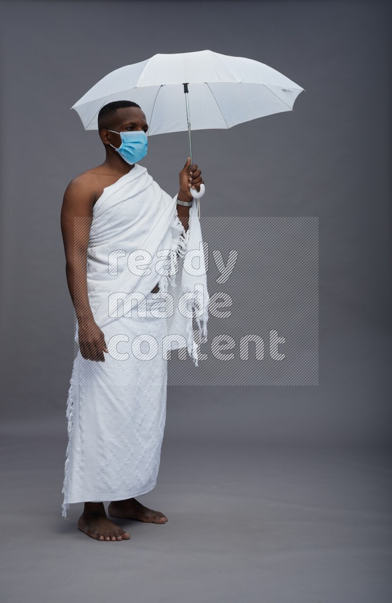 A man wearing Ehram with face mask Standing holding umbrella on gray background