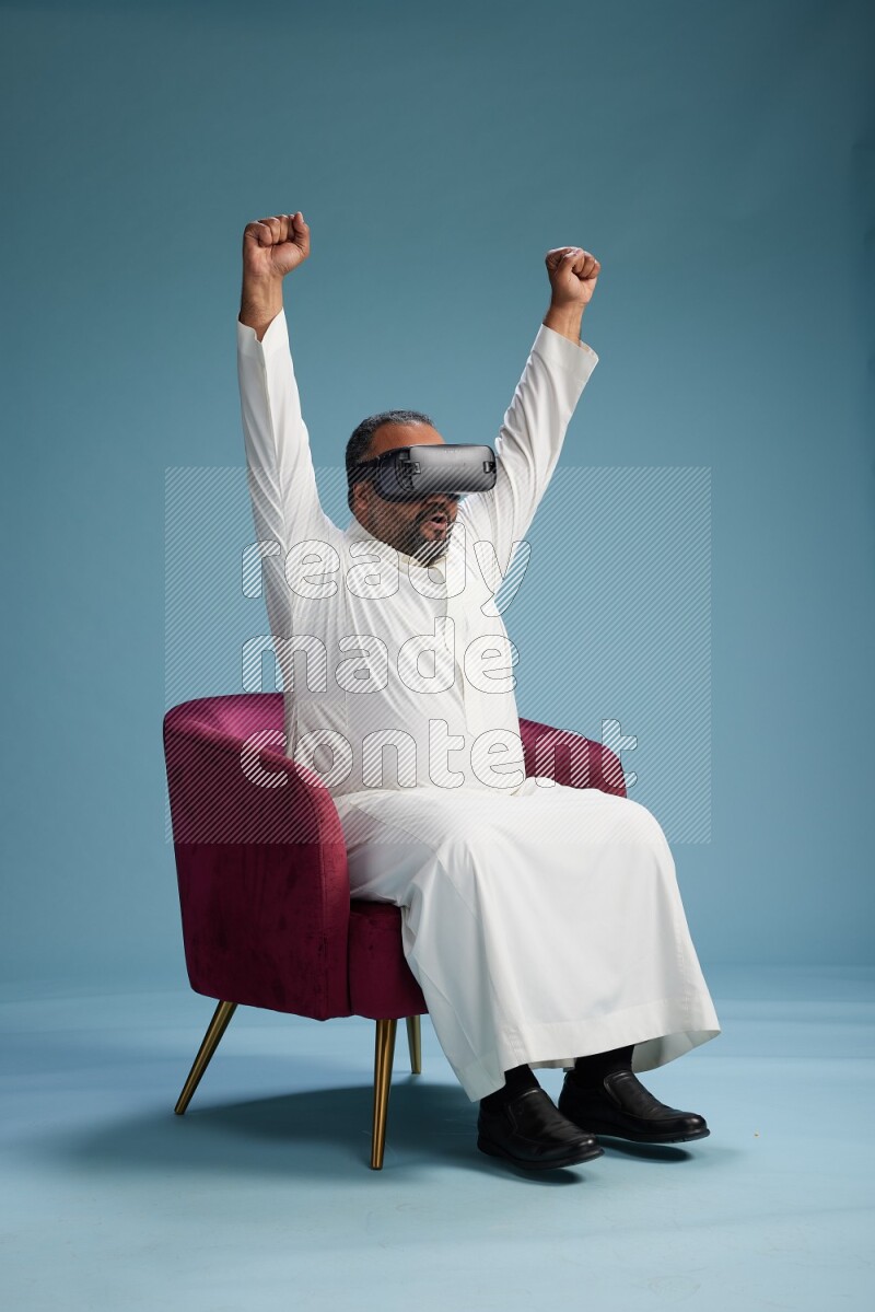 Saudi Man without shimag sitting on chair  gaming with VR on blue background