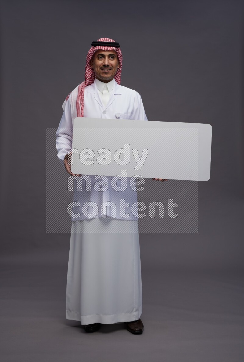Saudi man wearing thob with lab coat and shomag with pocket employee badge standing holding board on gray background