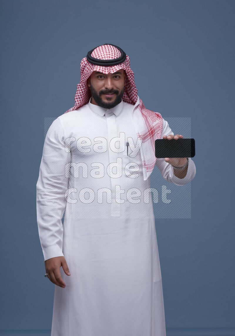 A man Showing his smartphone Screen on Blue Background wearing Saudi Thob