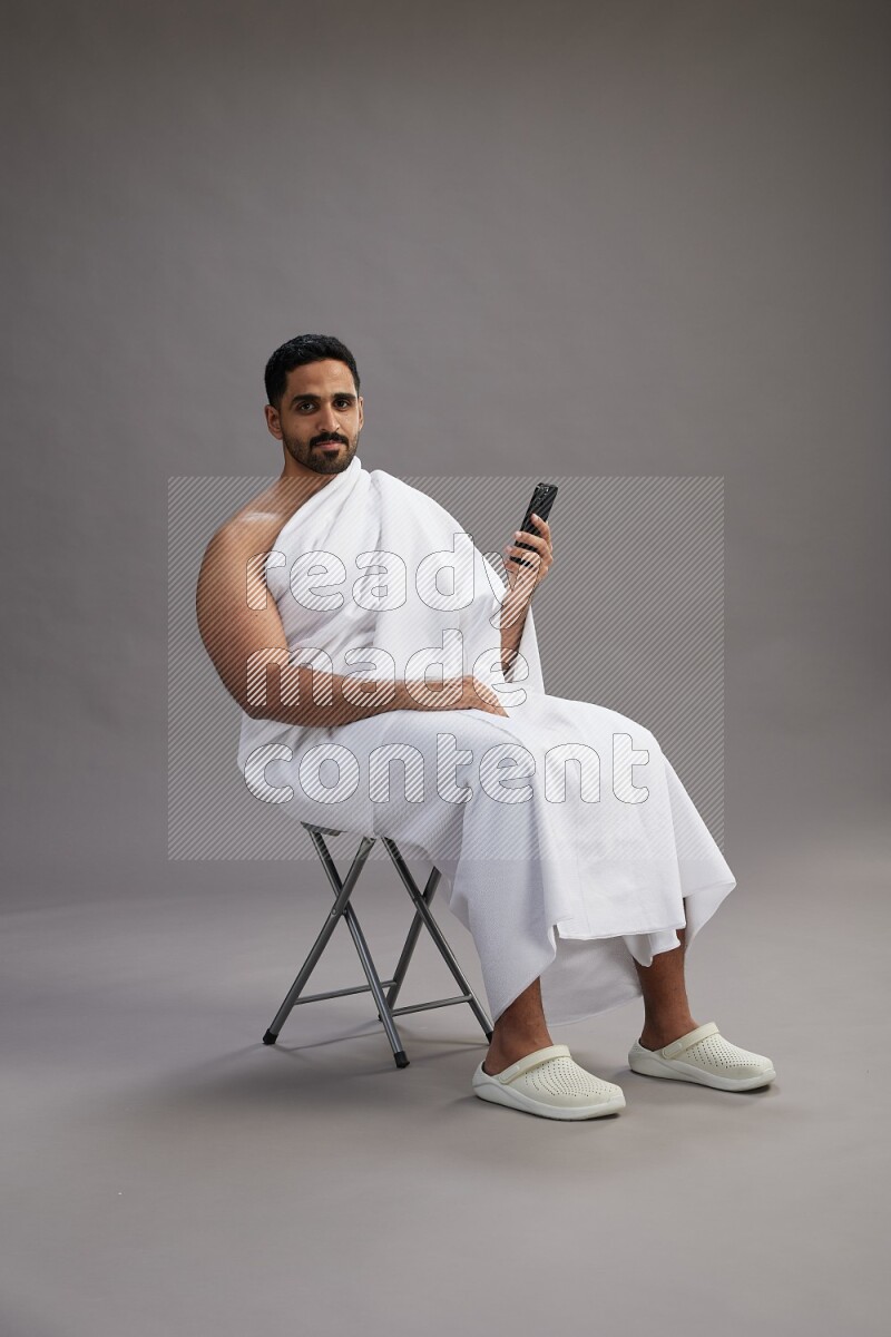 A man wearing Ehram sitting on chair texting on phone on gray background