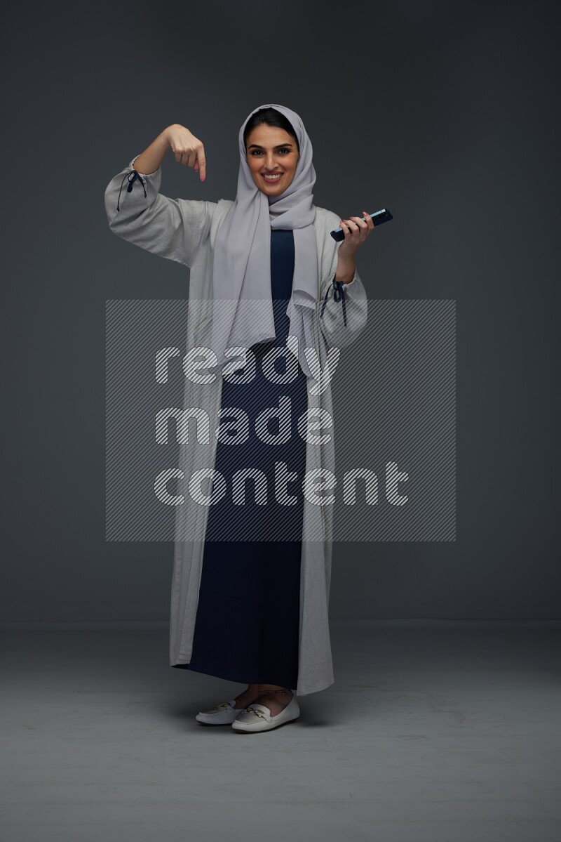A Saudi woman wearing a light gray Abaya and head scarf standing and holding a phone on a grey background