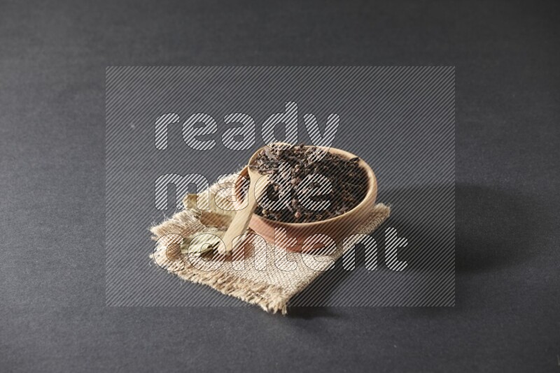 A wooden bowl, a wooden spoon full of cloves, and bay leaves (laurel) on a piece of burlap on a black flooring