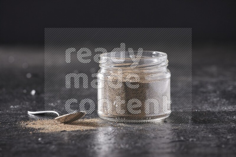 A glass jar full of black pepper powder and a metal spoon full of powder on a textured black flooring
