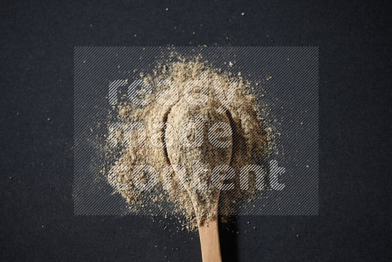 A wooden spoon full of cardamom powder and powder spreaded on black flooring