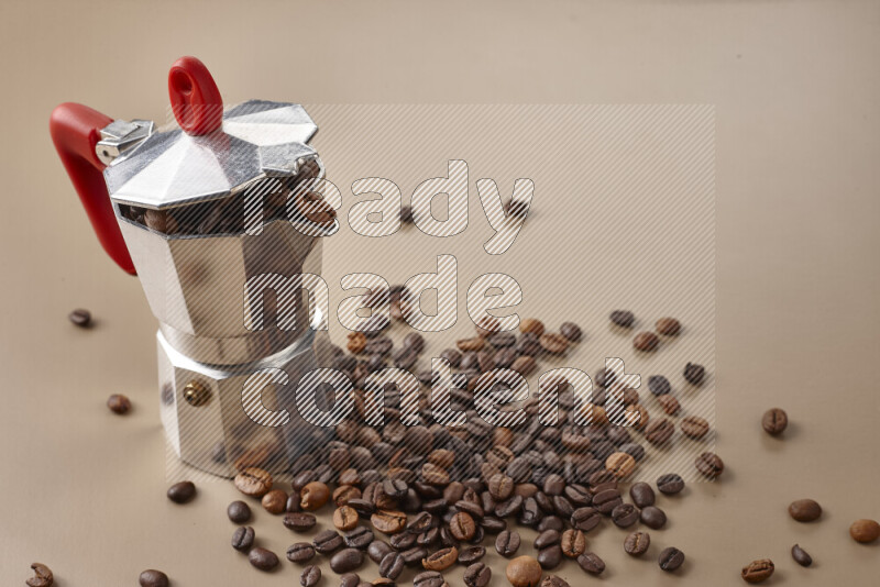 A moka pot with red handle surrounded by roasted coffee beans on beige background