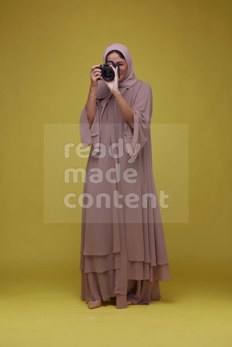 A woman Holding a Camera on a Yellow Background wearing Brown Abaya with Hijab