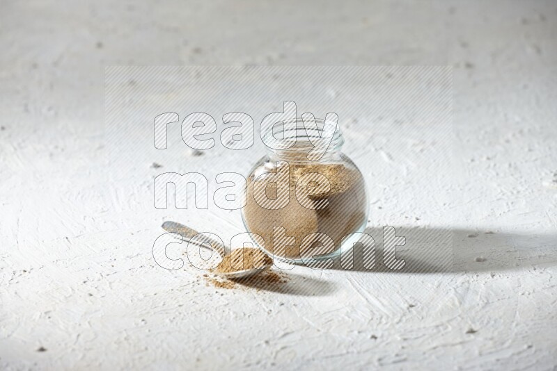 A glass spice jar and metal spoon full of cumin powder on textured white flooring