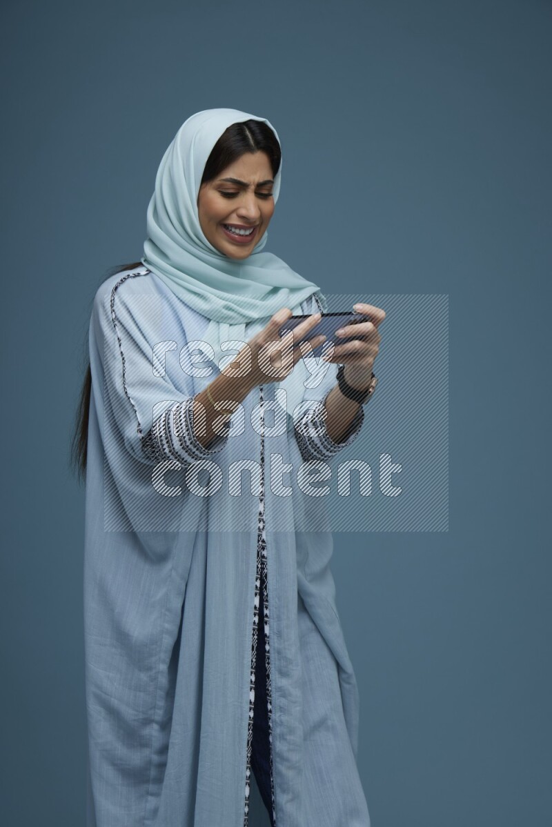 A Saudi woman Playing a Game on her phone on a blue background wearing a blue Abaya with hijab