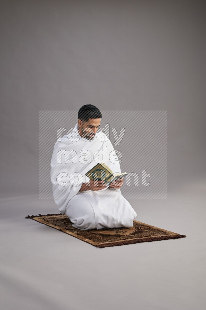 A man wearing Ehram sitting on floor reading quran on gray background