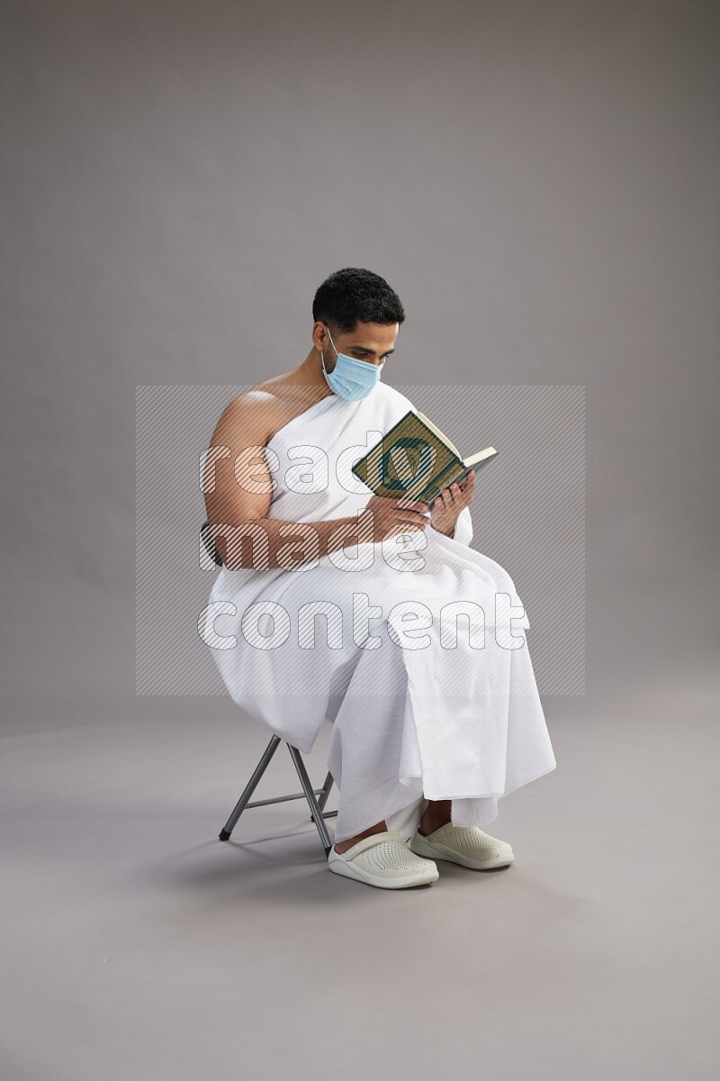 A man wearing Ehram with face mask sitting on chair reading quran on gray background