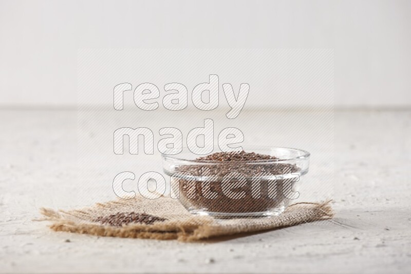 A glass bowl full of flax seeds and a bunch of seeds on burlap fabric on a textured white flooring