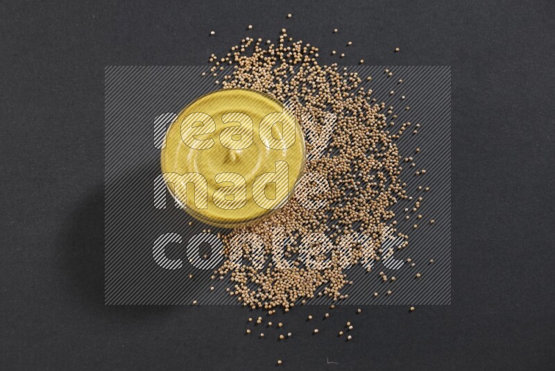 A glass bowl full of mustard paste with mustard seeds underneath on black flooring