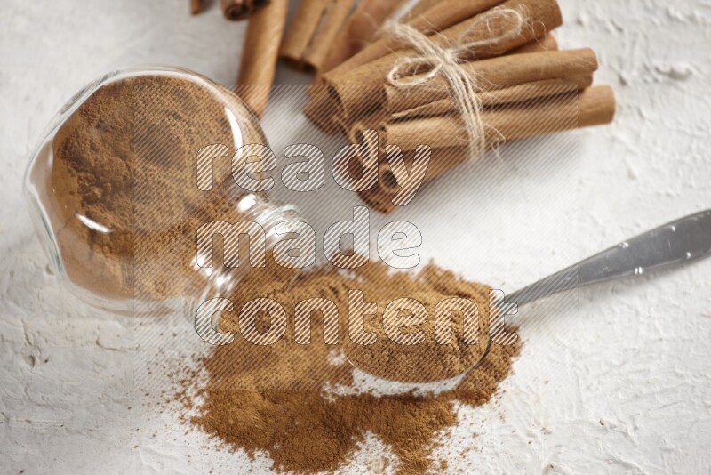 Flipped herbs glass jar full of cinnamon powder with a metal spoon full of powder and cinnamon sticks on a textured white background