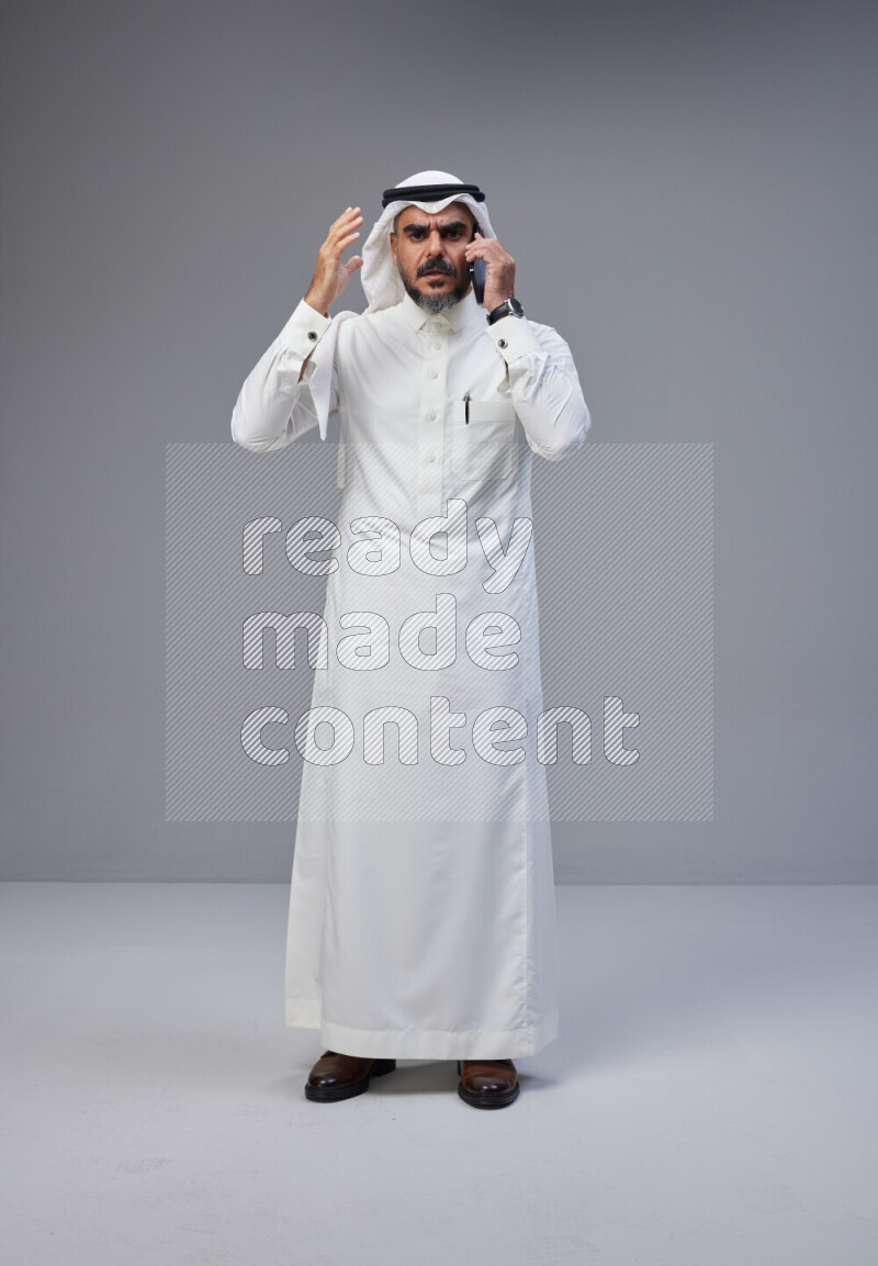Saudi man Wearing Thob and white Shomag standing talking on phone on Gray background
