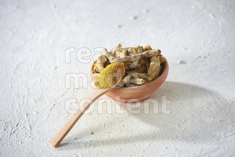 A wooden spoon full of turmeric powder above a wooden bowl full of dried turmeric whole fingers on a textured white flooring