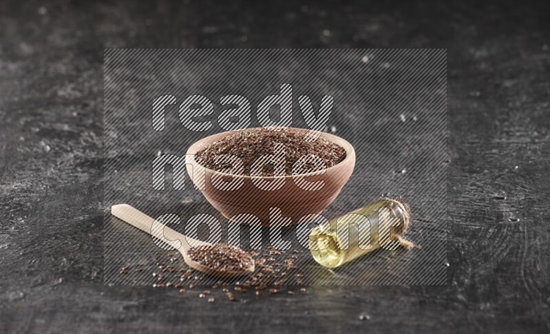 A wooden bowl and spoon full of flaxseeds and a glass bottle of flaxseeds oil on a textured black flooring