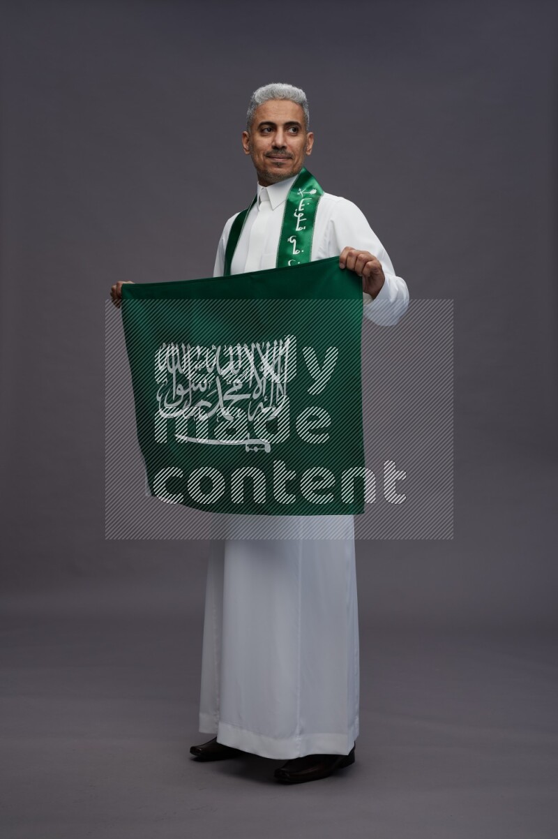 Saudi man wearing thob standing holding Saudi flag on gray background