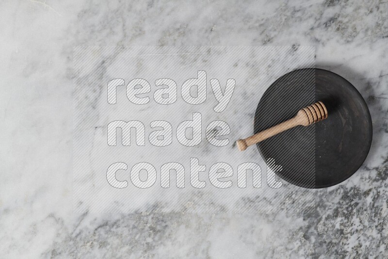 Black pottery plate with wooden honey dipper in it on grey marble background