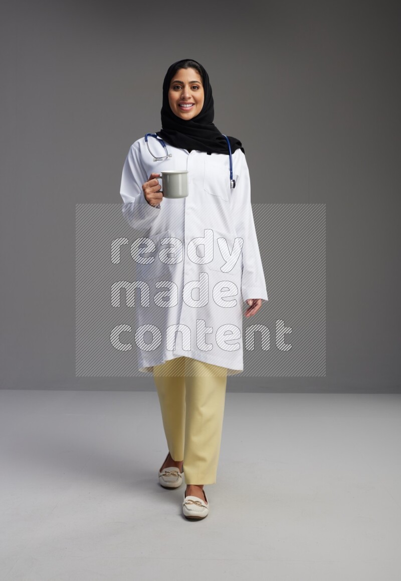 Saudi woman wearing lab coat with stethoscope standing holding mug on Gray background