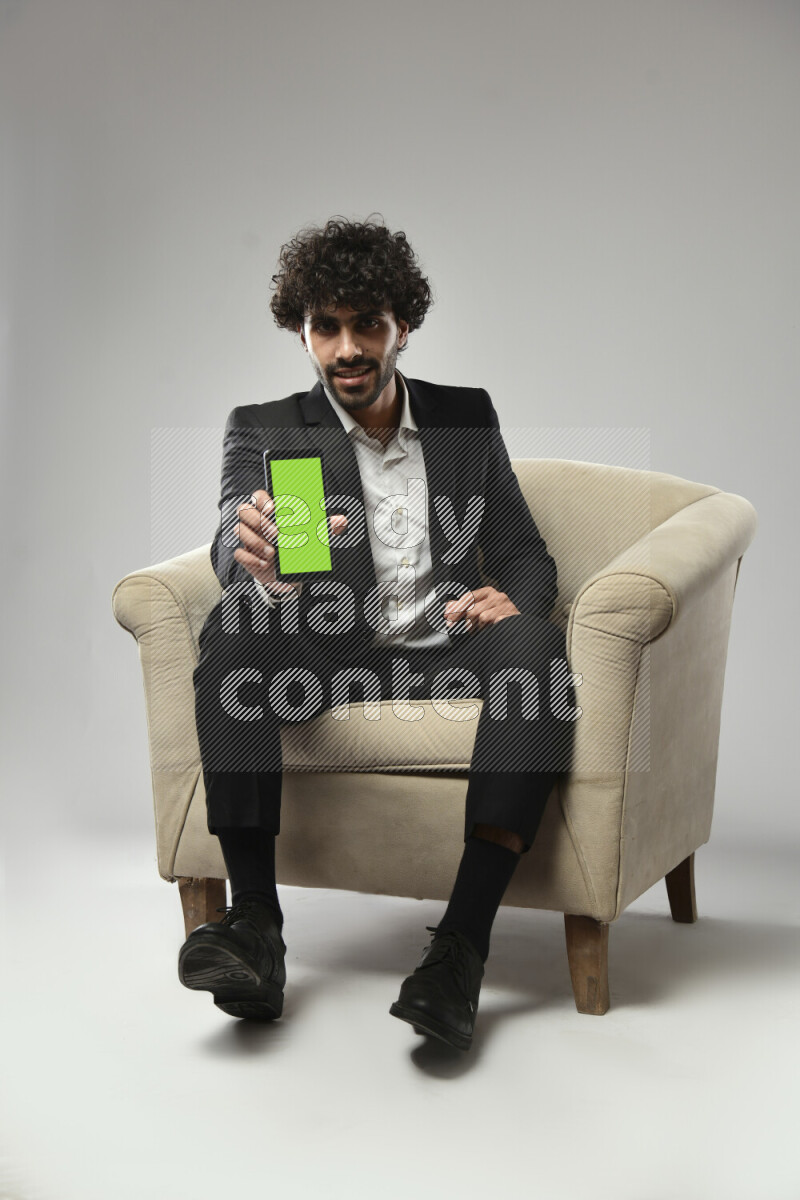 A man wearing formal sitting on a chair showing a phone screen on white background