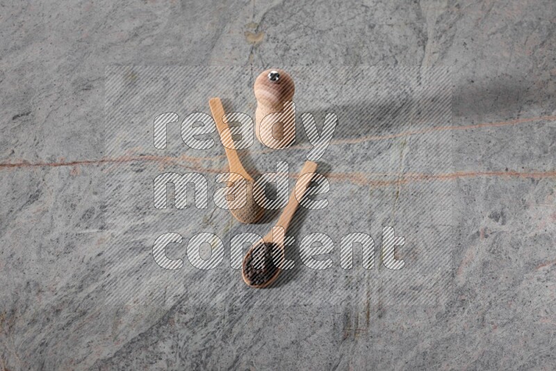 2 wooden spoons, one full of black pepper powder and the other full of black pepper beads and wooden grinder on a marble flooring