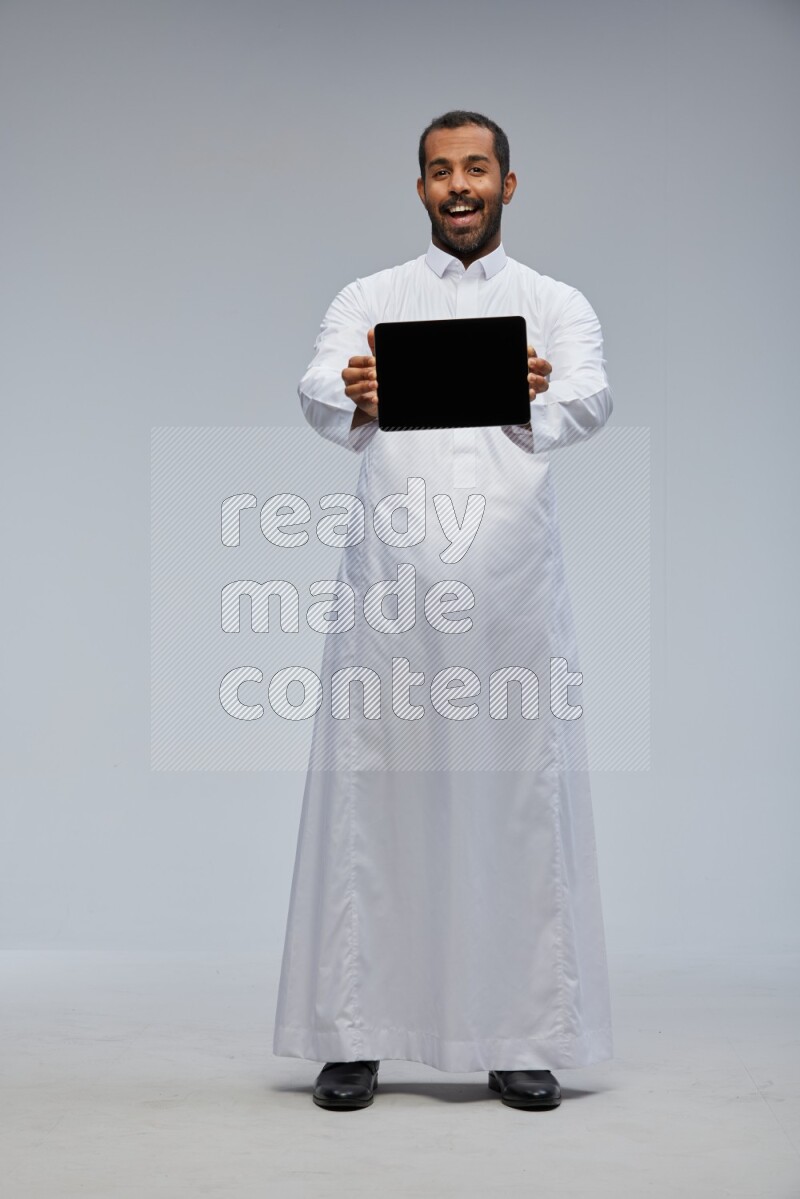 Saudi man wearing Thob standing showing tablet to camera on Gray background