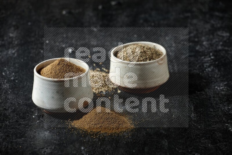 2 beige bowls, one full of cumin powder and the other full of seeds on a textured black flooring