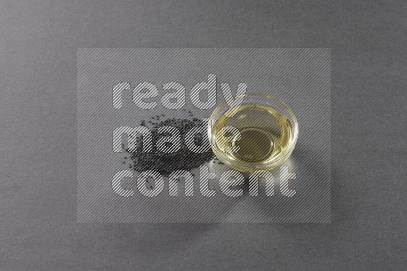 A glass bowl full of black seeds oil and black seeds beside it on a black flooring